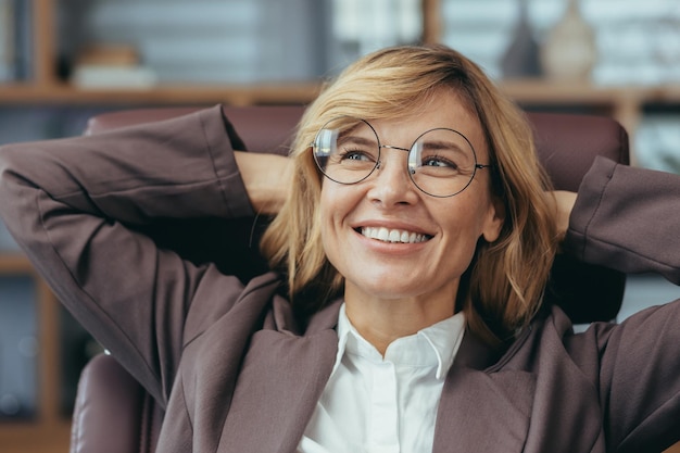 Successful blonde senior businesswoman with glasses relaxing and smiling in her stylish home office