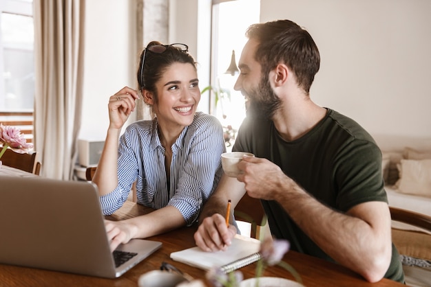 successful brunette couple man and woman drinking coffee and working on laptop together while sitting at table at home