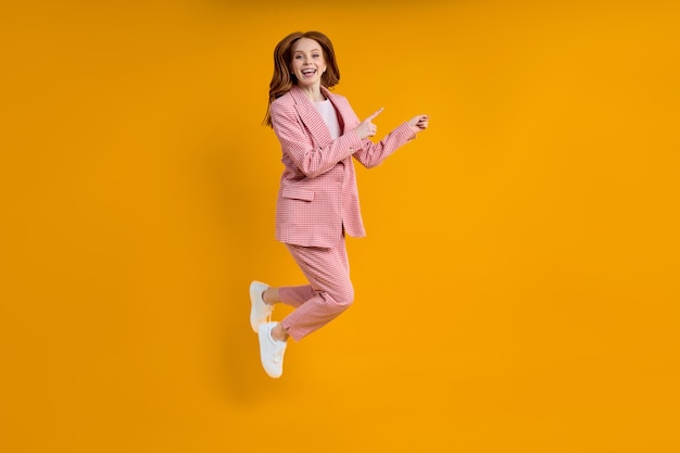 Successful business female in pink suit jumping pinting index finger at side, isolated on yellow background, shopping. portrait. copy space for advertisement