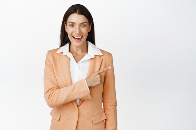 Successful business woman pointing finger right looking excited and smiling standing in suit against white background