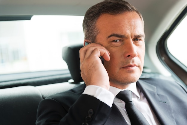 Successful businessman in car. Confident mature businessman talking on the mobile phone and looking away while sitting on the back seat of a car