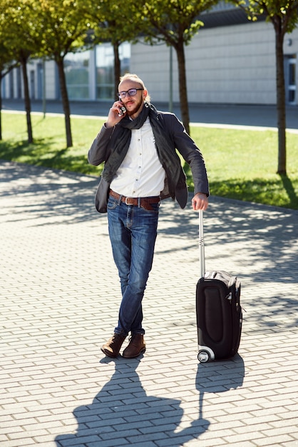 Photo successful businessman in casual suit pulls a suitcase, hurries to the airport and talks on smartphone. young caucasian man going on a business trip.