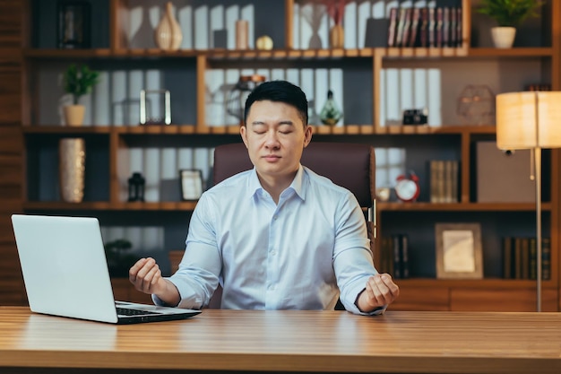 Successful businessman meditates sitting at a table in a classic office Asian man in shirt relaxes after working on laptop man at work