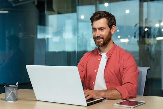 Successful businessman in red shirt happily working with laptop inside office mature man with beard