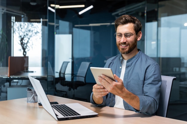 Successful businessman in a shirt uses a tablet computer a man sits at a desk smiling and happy a