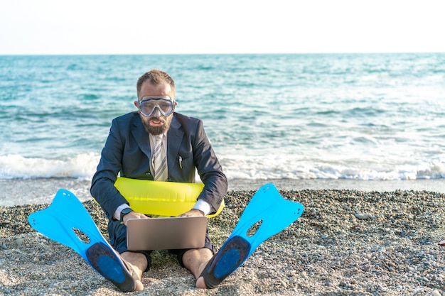 Successful businessman wearing snorkeling tools is working on the beach with laptop