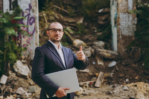 Successful businessman in white shirt, classic suit, glasses. Man show thumb up, stand with laptop pc computer phone near ruins, debris, stone building outdoors. Mobile Office, business, work concept.