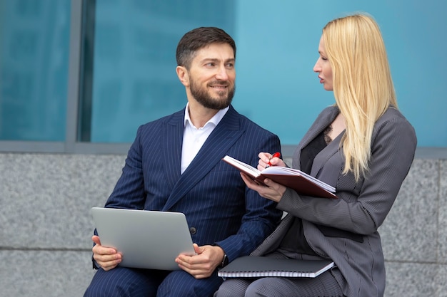 Successful businessmen man and woman are sitting on the stairs of a business building with documents and a laptop in their hands discussing business projects