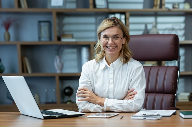 Successful businesswoman boss in shirt sitting at table with crossed arms smiling and looking at