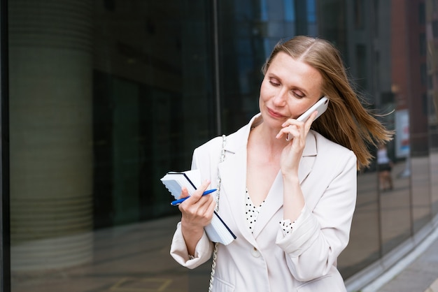 Successful businesswoman or businesswoman taking notes and talking on cell phone while walking outdo...