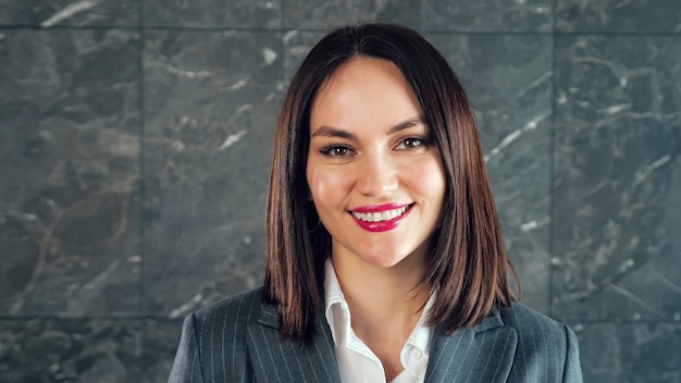 Successful businesswoman with red lipstick smiles posing for camera against grey marble wall with white patterns in office close view