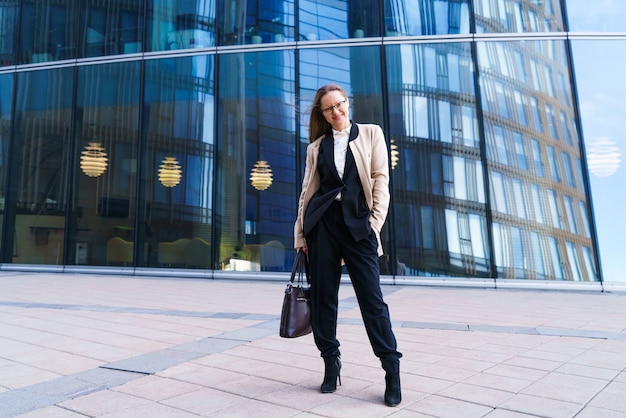 A successful caucasian woman in a coat and suit and glasses stands in front of an office building wi...