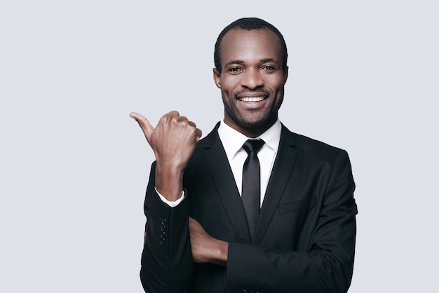 Successful choice. Handsome young African man in formalwear pointing away and smiling while standing against grey background