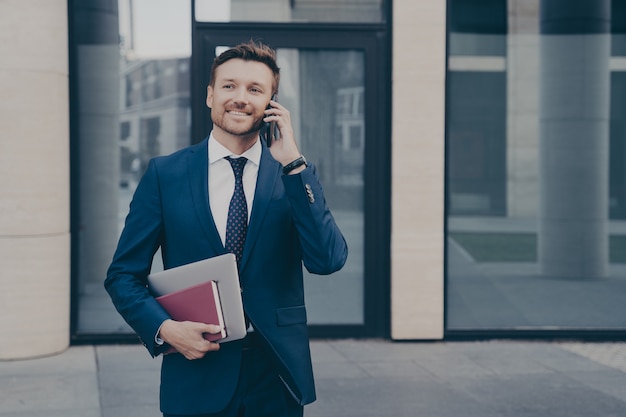 Successful and confident business owner in formal wear, talking on phone with his employee, telling directions and instructions on next move, holding note book with laptop, blurred background