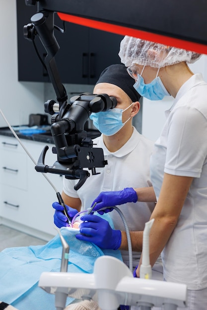 A successful dentist looks at the patients teeth with a dental microscope and holds dental instruments near his mouth The assistant helps the doctor They wear white uniforms with masks and gloves