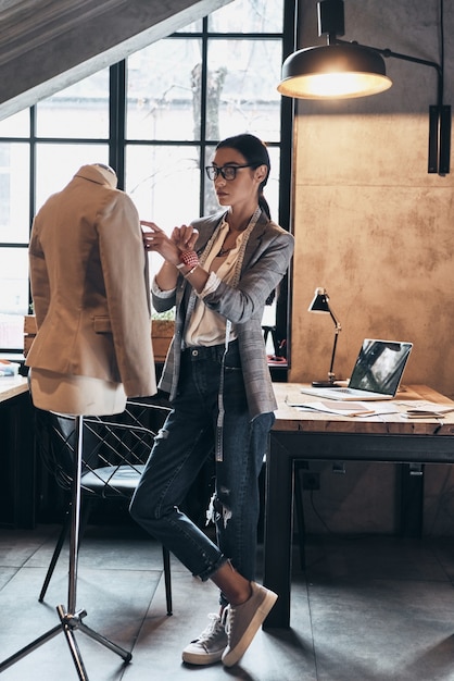 Photo successful fashion designer. full length of serious young woman in eyewear using sewing needles for sewing a jacket on mannequin while standing in her workshop