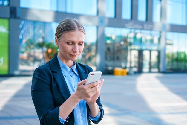 Successful female banker using smart phone outdoors while standing near office