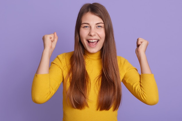 Successful female student looks happy, clenches fists, accomplishes goal, exclaims finally victory, stands amused isolated over purple wall.