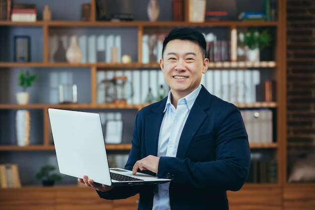 Successful and happy businessman standing working on laptop smiling and looking at camera Asian working in a classic home office for dinner