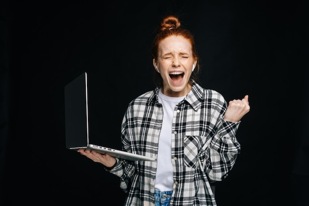 Successful happy shocked young woman with open mouth and closed eyes holding laptop computer