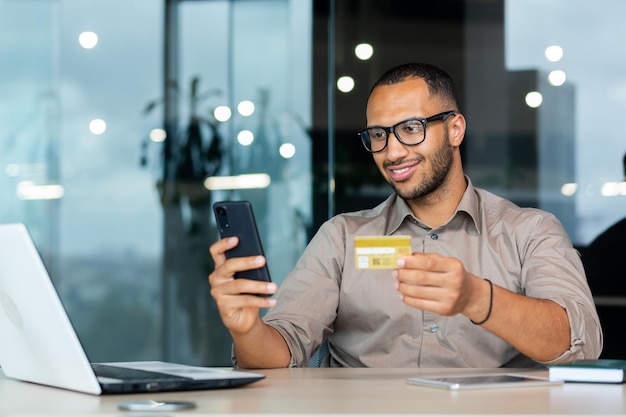 Successful hispanic businessman inside office man in shirt smiling and happy holding bank credit