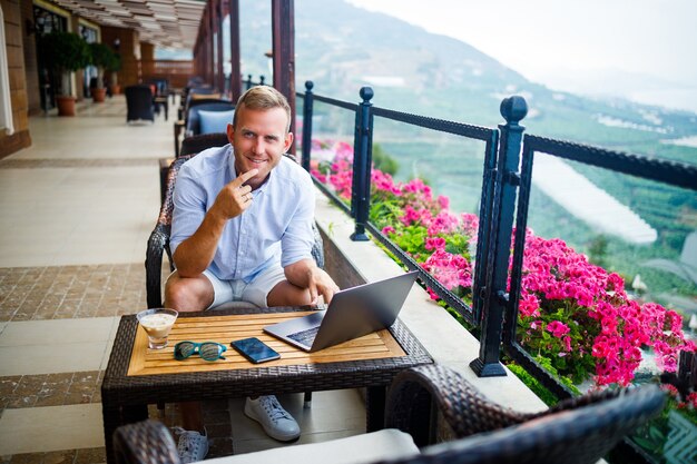 A successful male businessman is sitting at a table with a laptop, he is working on vacation. Vacation and remote work. The guy is sitting in a restaurant on a terrace with a panoramic view