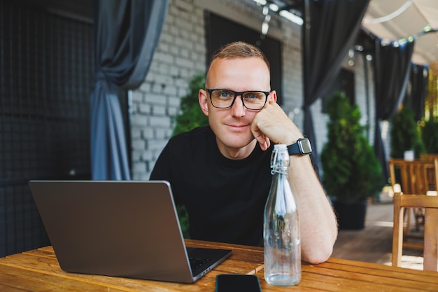 A successful male freelancer in a black tshirt and glasses sits on the summer terrace of a cafe and works on a laptop The manager sits in a cafe during lunch and works online