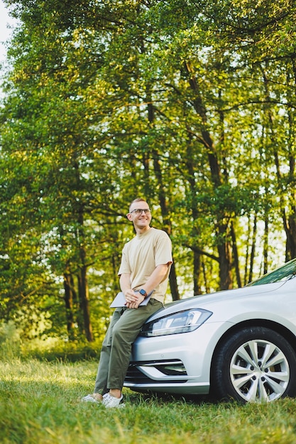 A successful man in glasses works on a laptop while leaning on the hood of his car Remote work in nature Work on a laptop online Working on a computer while traveling