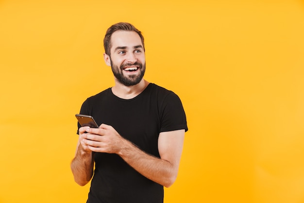 successful man wearing basic black t-shirt smiling and holding smartphone isolated over yellow wall