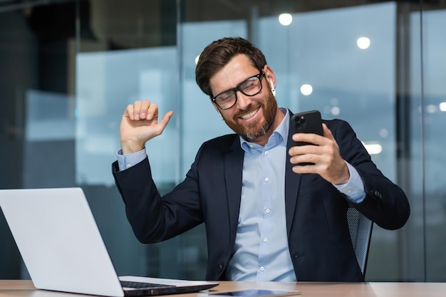 Successful mature businessman in office listening to music and dancing boss in glasses with phone