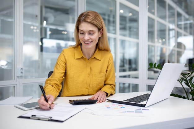 Photo successful mature female financial accountant at workplace inside office fills data in table works