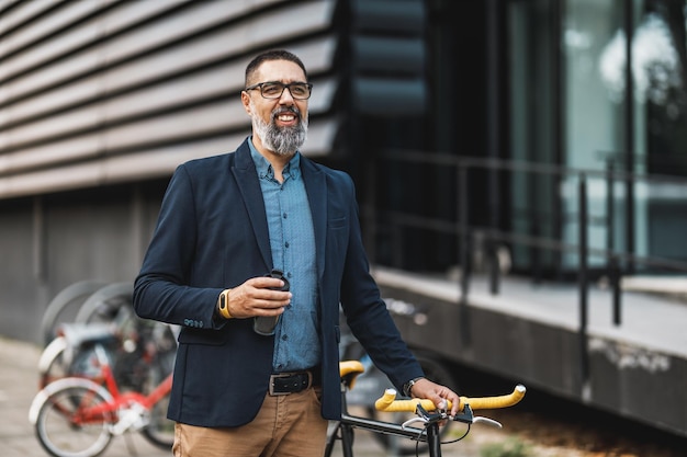 Photo successful middle-aged businessman with a cup of coffee to go and with bicycle beside him, walks in front of office district.