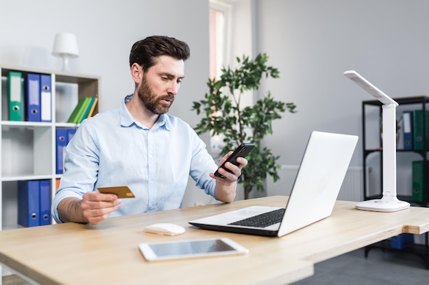 Successful online shopping through a smartphone Successful deal Satisfied young man holds a credit card and a mobile phone makes an order Sitting at a desk in the office on a laptop