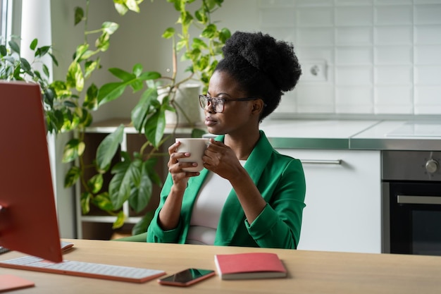 Successful pensive African American woman drinking coffee sits at table with computer in home office