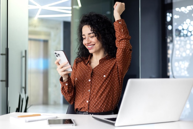 Successful satisfied business woman celebrating success and triumph boss at workplace holding phone