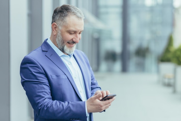 Successful senior and experienced grayhaired man outside office building reading message on phone