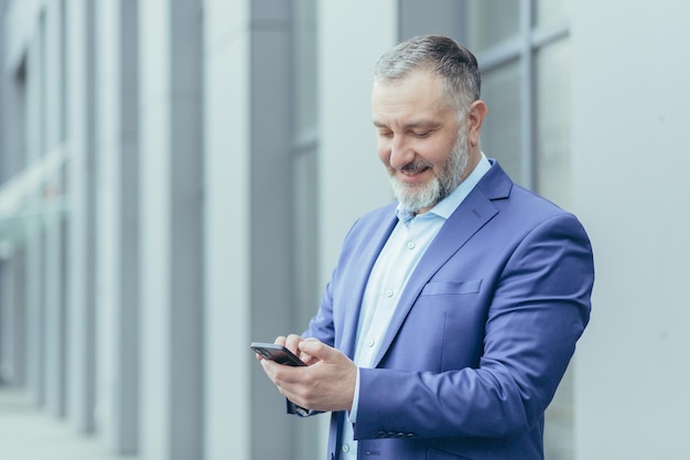 Successful senior and experienced grayhaired man outside office building reading message on phone