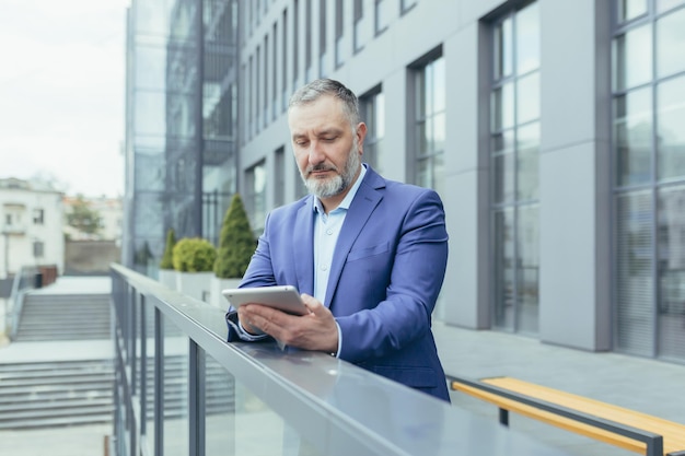 Successful and serious pensive senior gray haired man outside office building using tablet computer