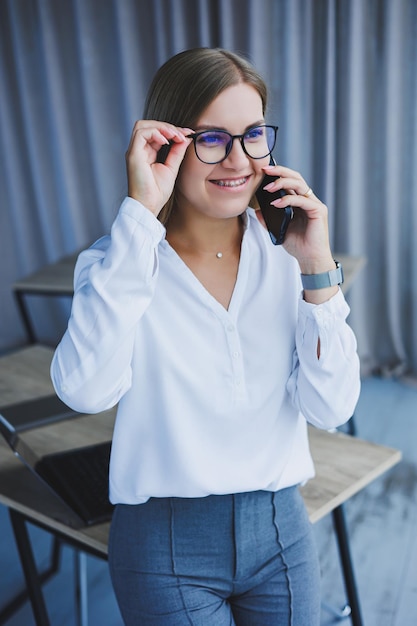 A successful smiling business woman is standing in the office by the window with a phone in her hands A female manager in glasses and a white shirt is talking on the phone