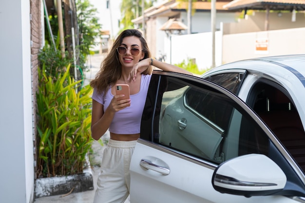 Successful smiling fit tanned attractive woman in casual wear is using her smart phone while standing near luxury modern car outdoorsxA