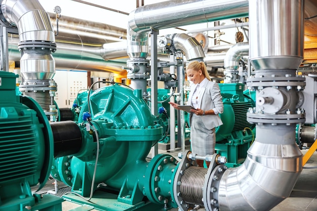 Successful smiling hardworking female manager in suit standing in heating plant with tablet in hands and checking on turbine