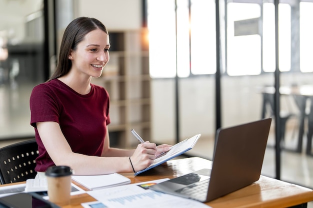 Successful smiling young businesswoman using laptop and computer while doing some paperwork at the office