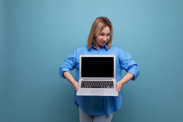 Successful smiling young woman in casual shirt showing laptop screen with mockup on blue isolated