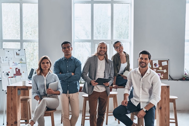 Successful team. Group of young modern people in smart casual wear smiling and looking at camera while standing in the creative office