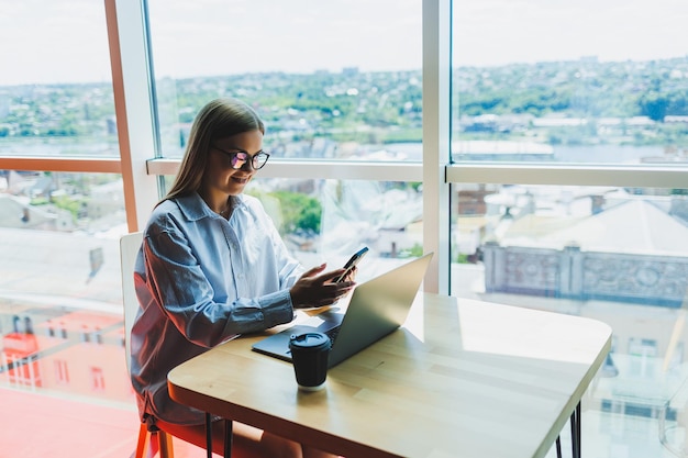 Successful woman is looking at a laptop in a cafe and drinking coffee A young smiling woman in glasses sits at a table near the window with a phone Freelance and remote work Modern female lifestyle