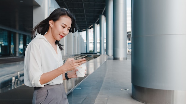 Successful young Asia businesswoman in fashion office clothes holding disposable paper cup of hot drink and using smart phone while walking outdoors in urban modern city