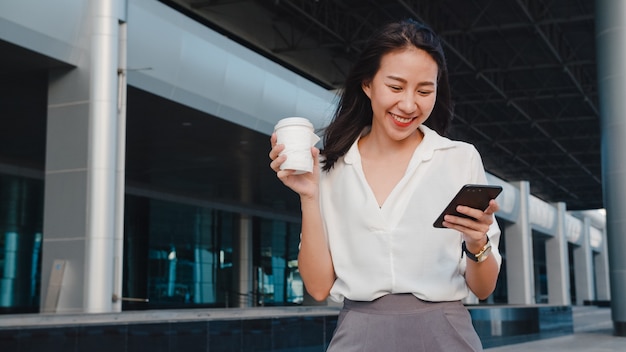 Successful young Asia businesswoman in fashion office clothes holding disposable paper cup of hot drink and using smart phone while walking outdoors in urban modern city