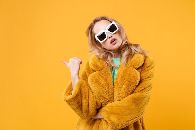 Successful young blonde woman girl in yellow fur coat, dark sunglasses posing isolated on orange wall background studio portrait. People lifestyle concept. Mock up copy space. Pointing thumb aside.