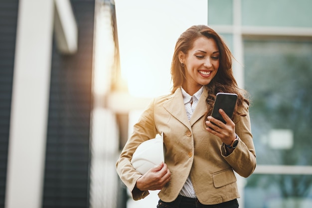 Successful young female engineer with white helmet checking social media on her smartphone and having a quick break in front of office district.