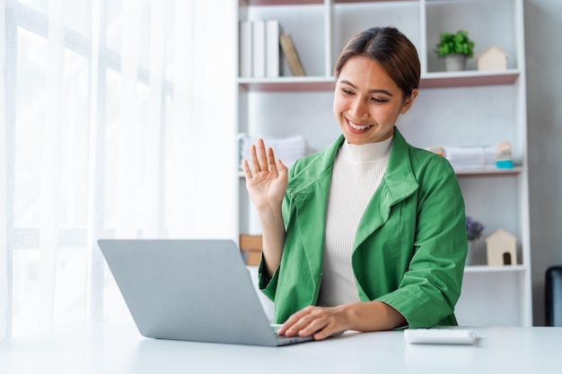 Successful young pretty Asian woman waving hello talking on a video call when using laptop computer at desk in office workplace